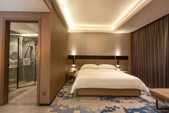 Habitación de hotel con cama, lámparas de noche, baño con ducha y un ventanal grande.
