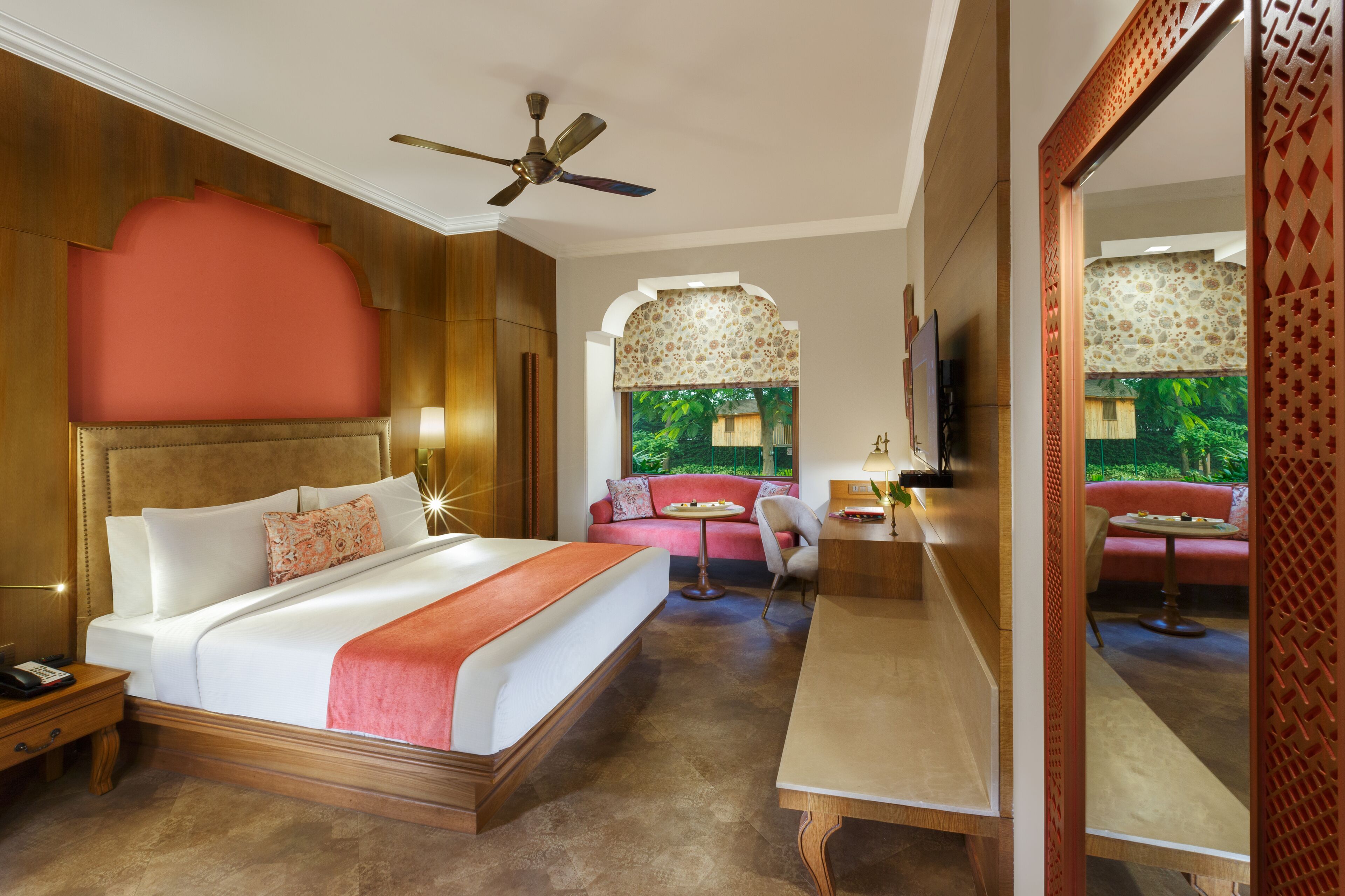 A hotel room with a large bed, a desk, a chair, a sofa, a ceiling fan, and a TV.