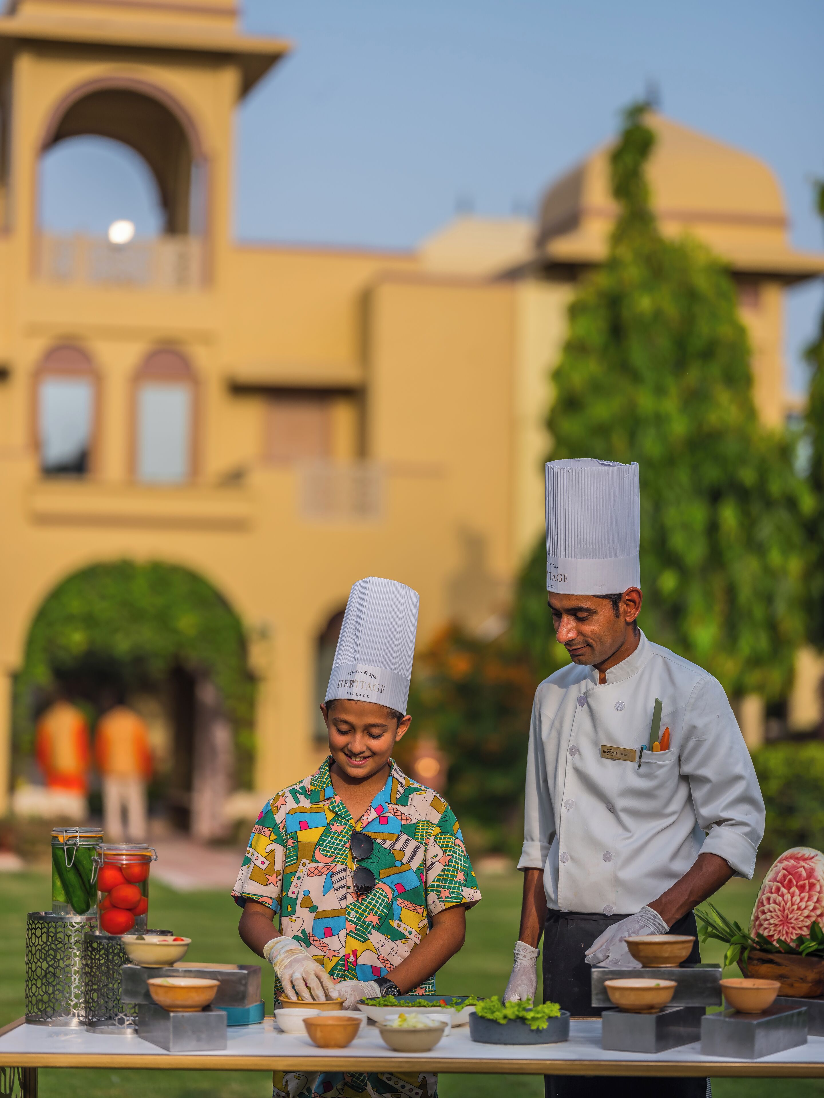Two chefs preparing food outdoors with a large building in the background.