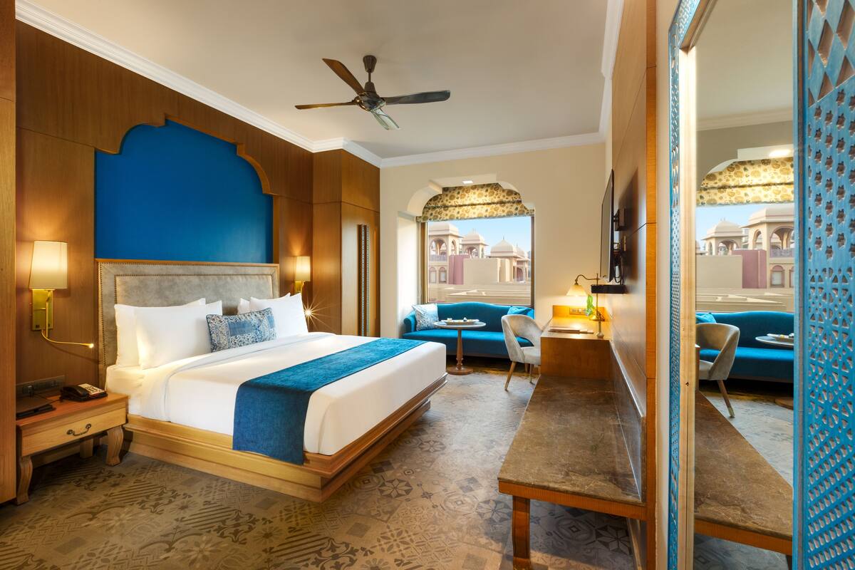 A hotel room with a large bed, a desk, a chair, and a sofa. There is a ceiling fan and a view of the outside through a window.