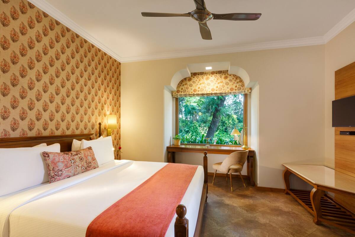 A hotel room with a bed, desk, chair, and a view of greenery through a window.
