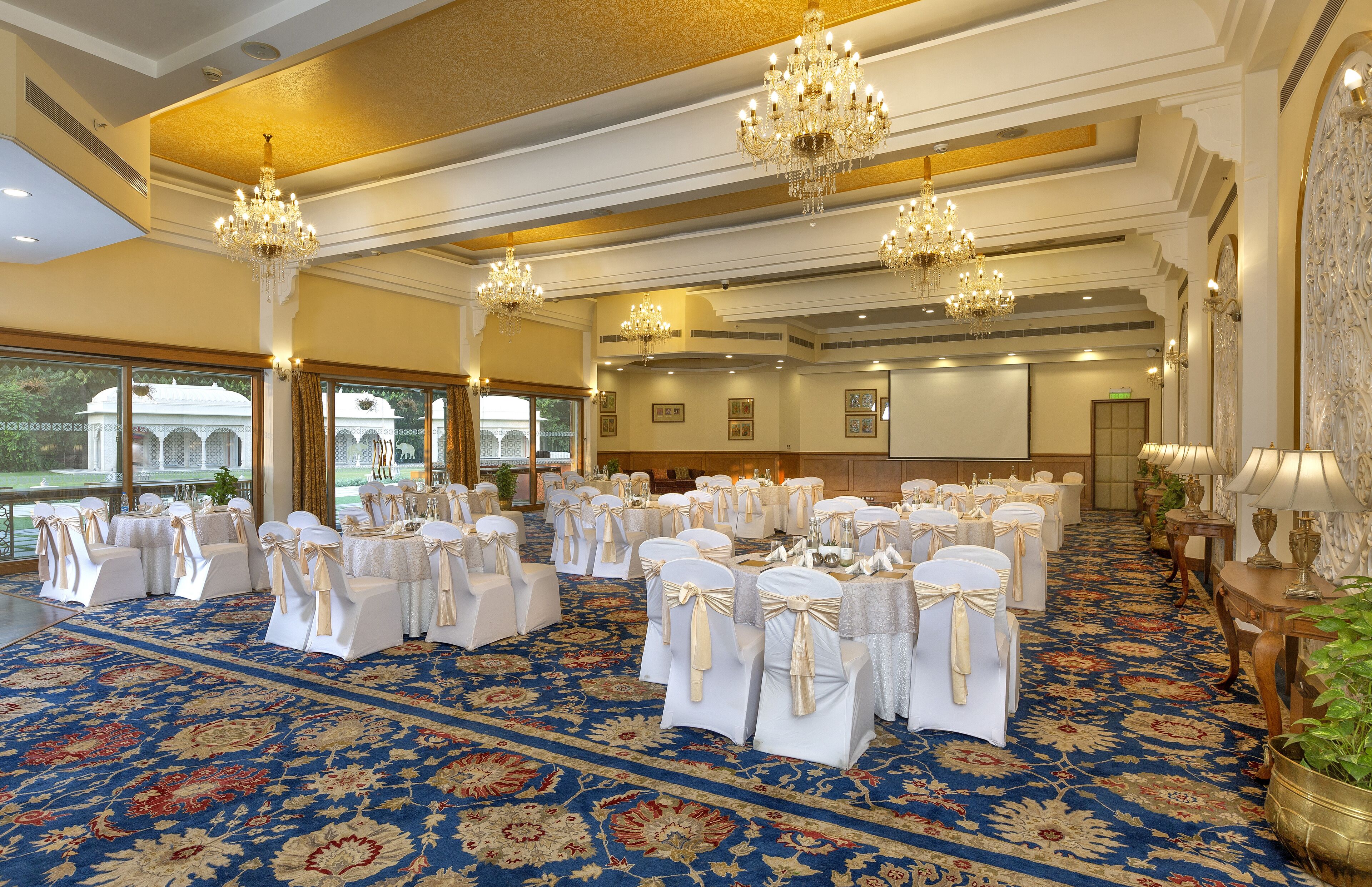A banquet hall with round tables, white chairs, chandeliers, and a patterned carpet.