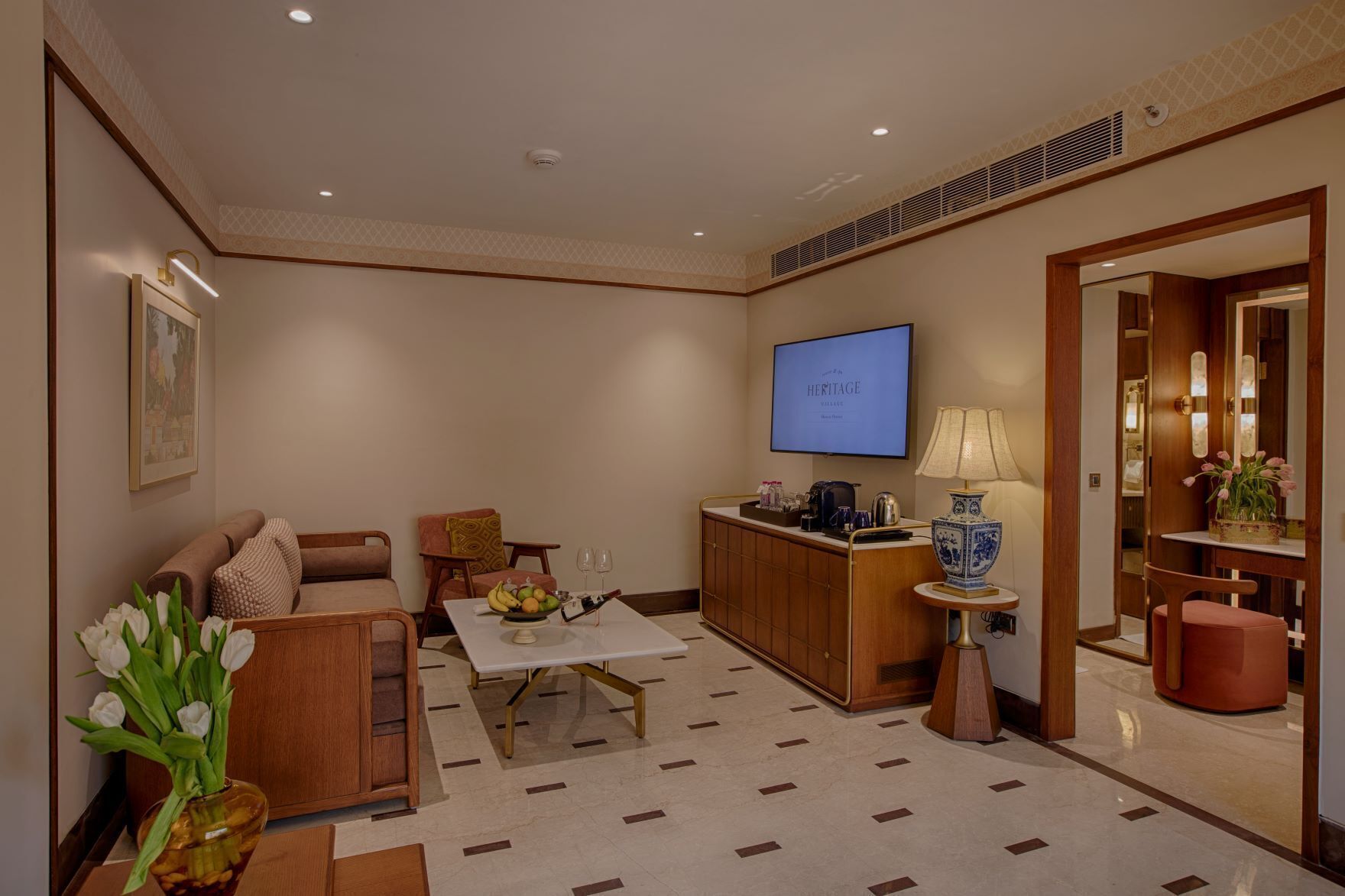 A modern hotel room with a sofa, a dining table, a small kitchen area, a TV, and a decorative vase.