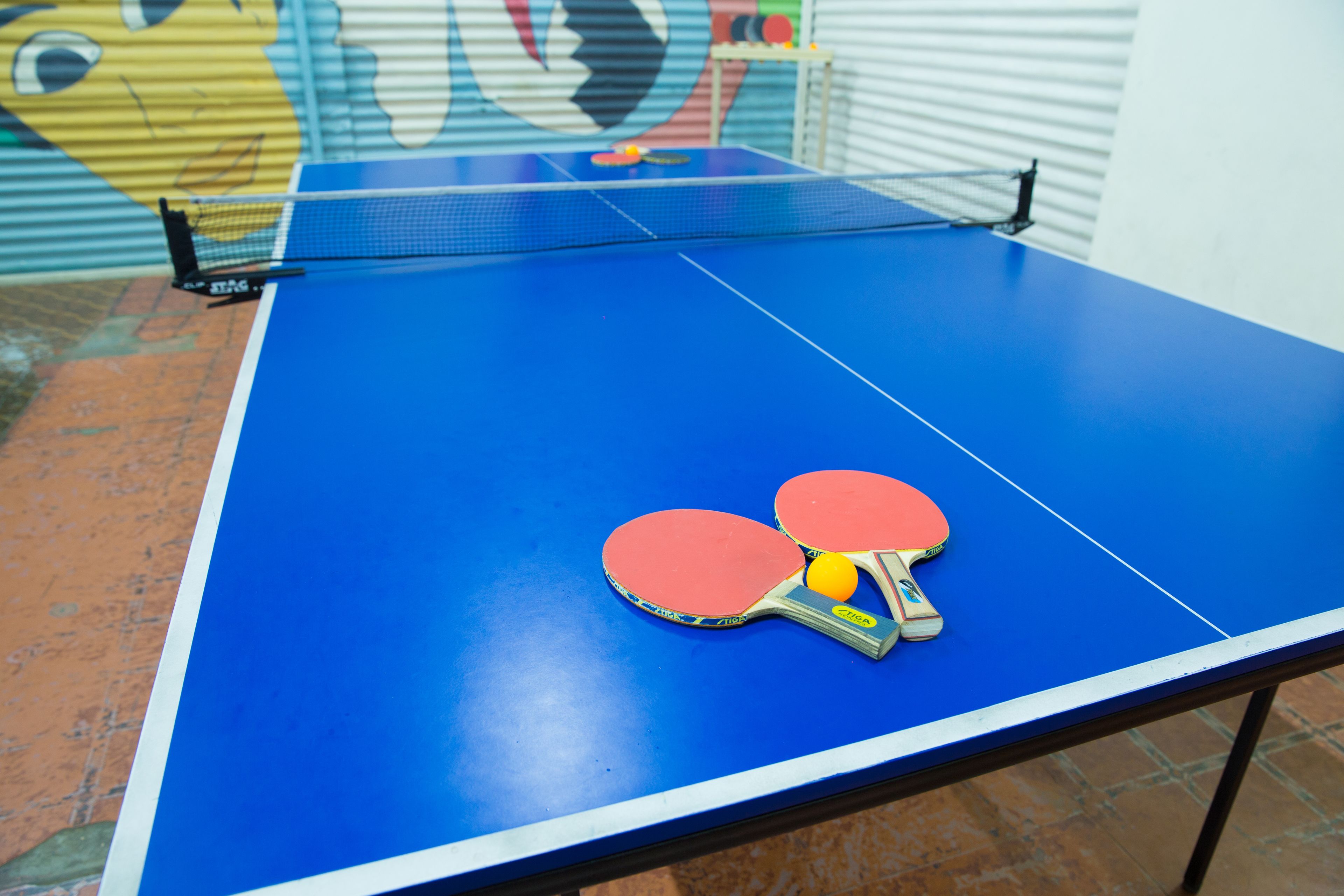 A blue ping pong table with two red paddles and a yellow ball.