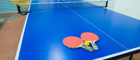 A blue ping pong table with two red paddles and a yellow ball.