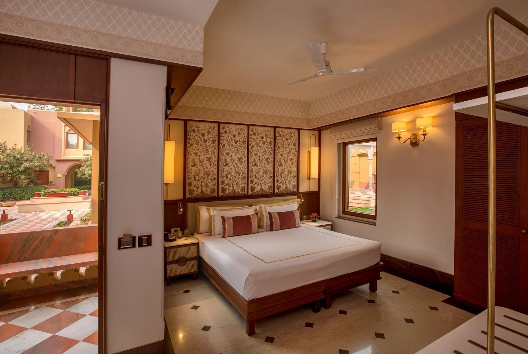 A hotel room with a bed, bedside table, lamp, and a view of the outside.
