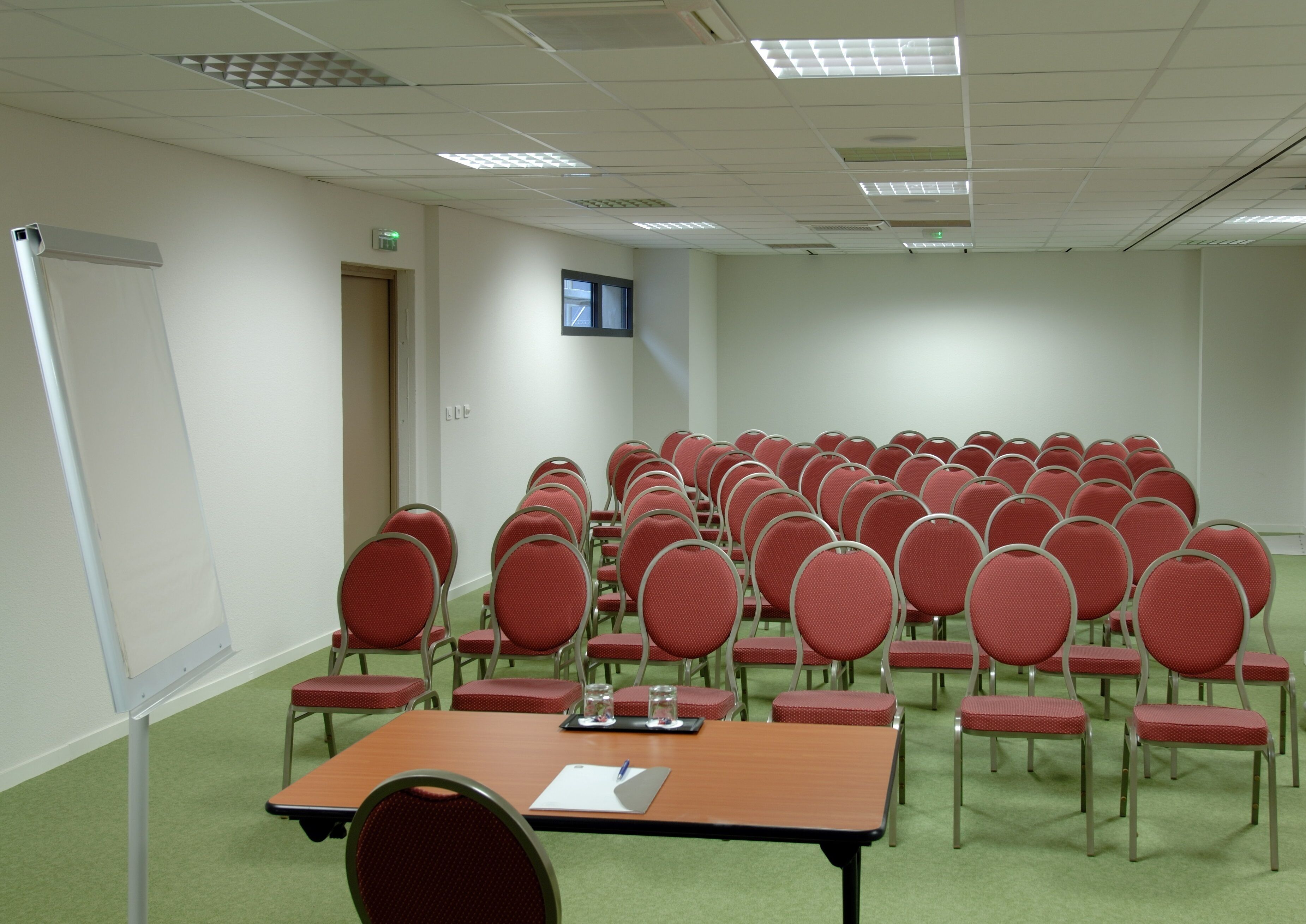 Un salón de conferencias con filas de sillas rojas, una pantalla de proyector y una mesa con un cuaderno y un bolígrafo.
