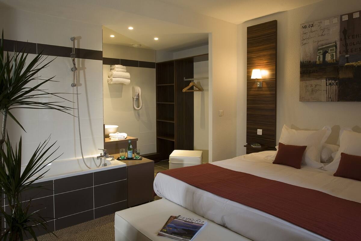 Una habitación de hotel moderna con una cama grande, un área de ducha separada, un pequeño rincón de descanso y un baño con bañera.