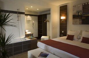 Una habitación de hotel moderna con una cama grande, un área de ducha separada, un pequeño rincón de descanso y un baño con bañera.