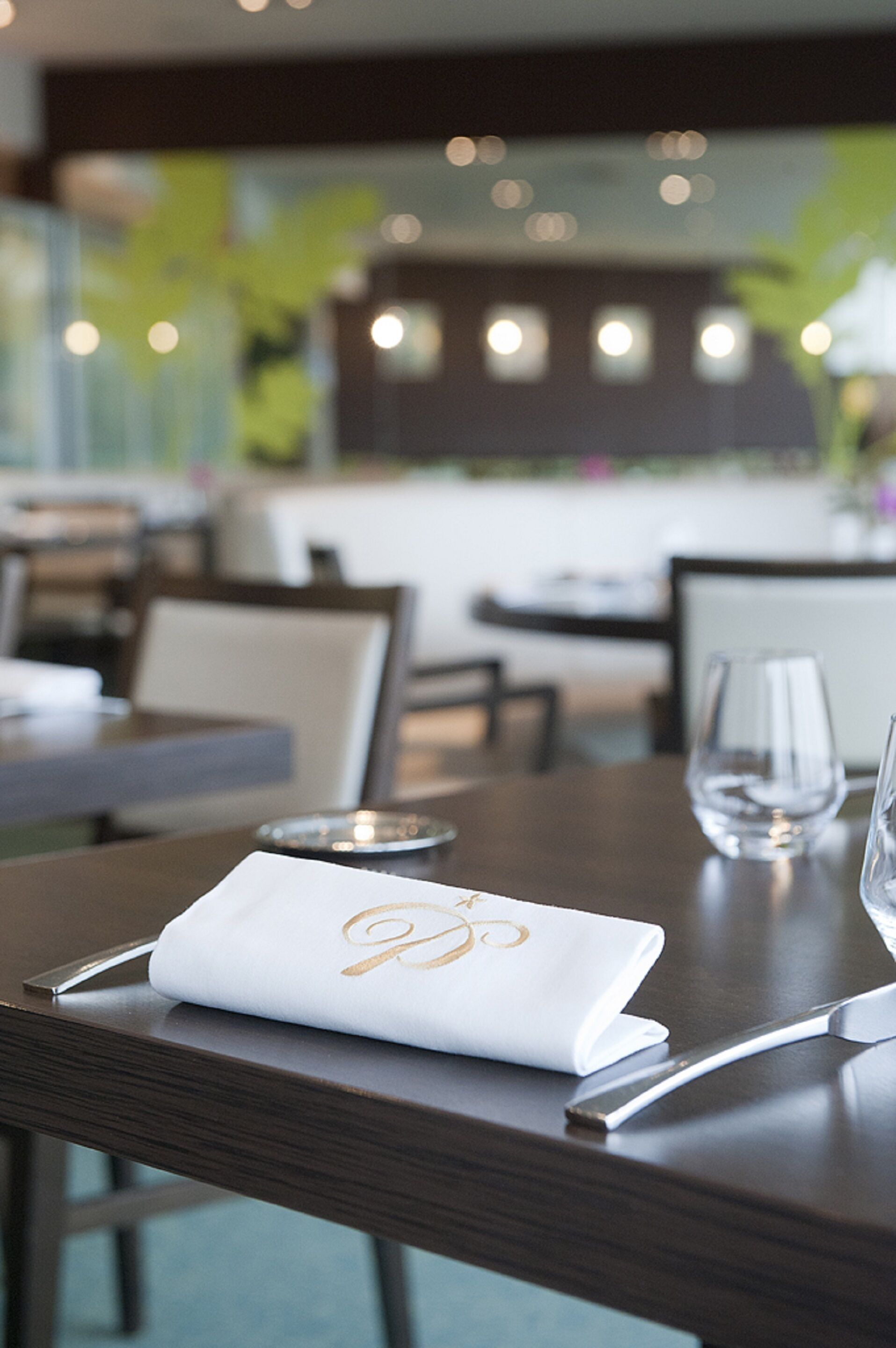 Ein Restaurantambiente mit gedecktem Tisch, darunter ein gefaltetes Monogramm-Serviet, ein Weinglas und eine Kerze.