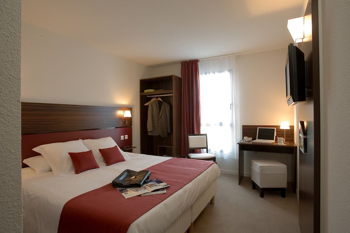 Habitación de hotel con una cama grande, un escritorio con computadora, televisión, armario y ventana con cortinas.