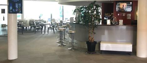 Zona de recepción de un hotel con mostrador, taburetes de bar, una planta en maceta y un área de estar.