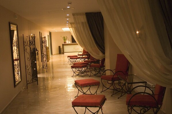 Um hall de hotel com um longo corredor, cadeiras estofadas vermelhas, um balcão de recepção e uma planta.