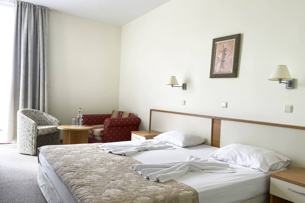 A hotel room with a large bed, a chair, a small table, and a wall-mounted lamp.