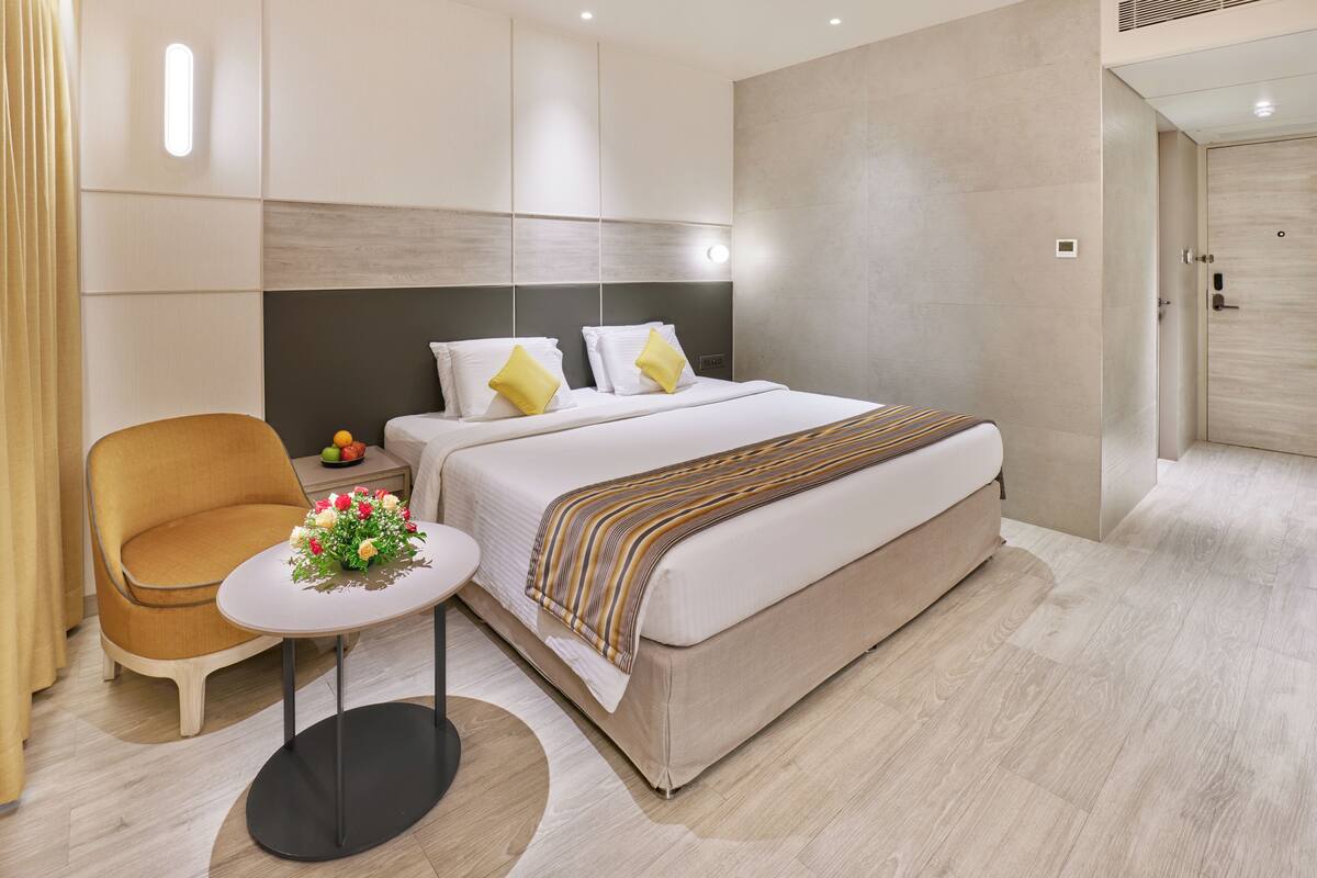 A modern hotel room with a bed, bedside table, chair, and a small table with flowers.