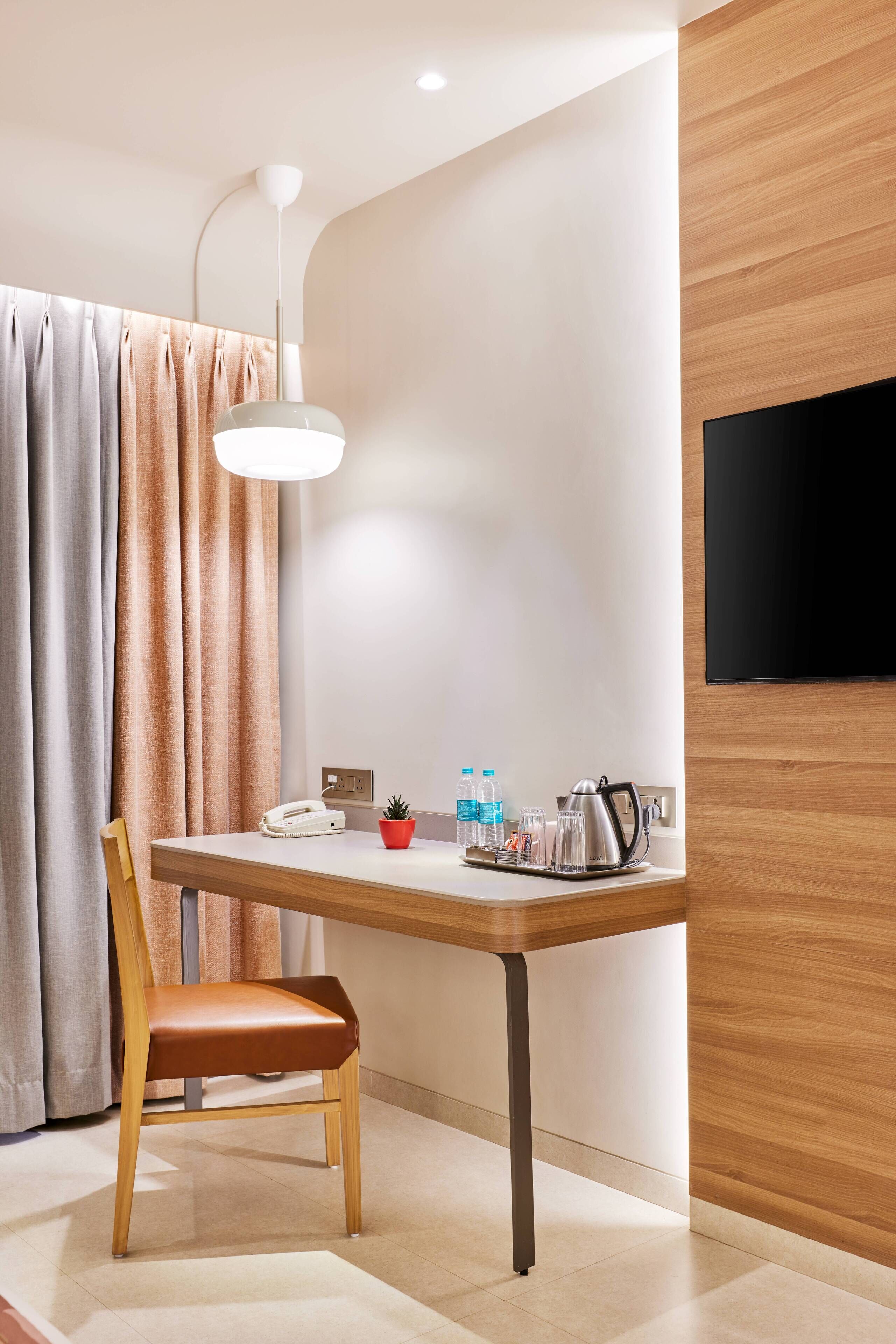 A modern hotel room with a wooden dining table, a single chair, a flat-screen TV mounted on the wall, and a small potted plant on the table.