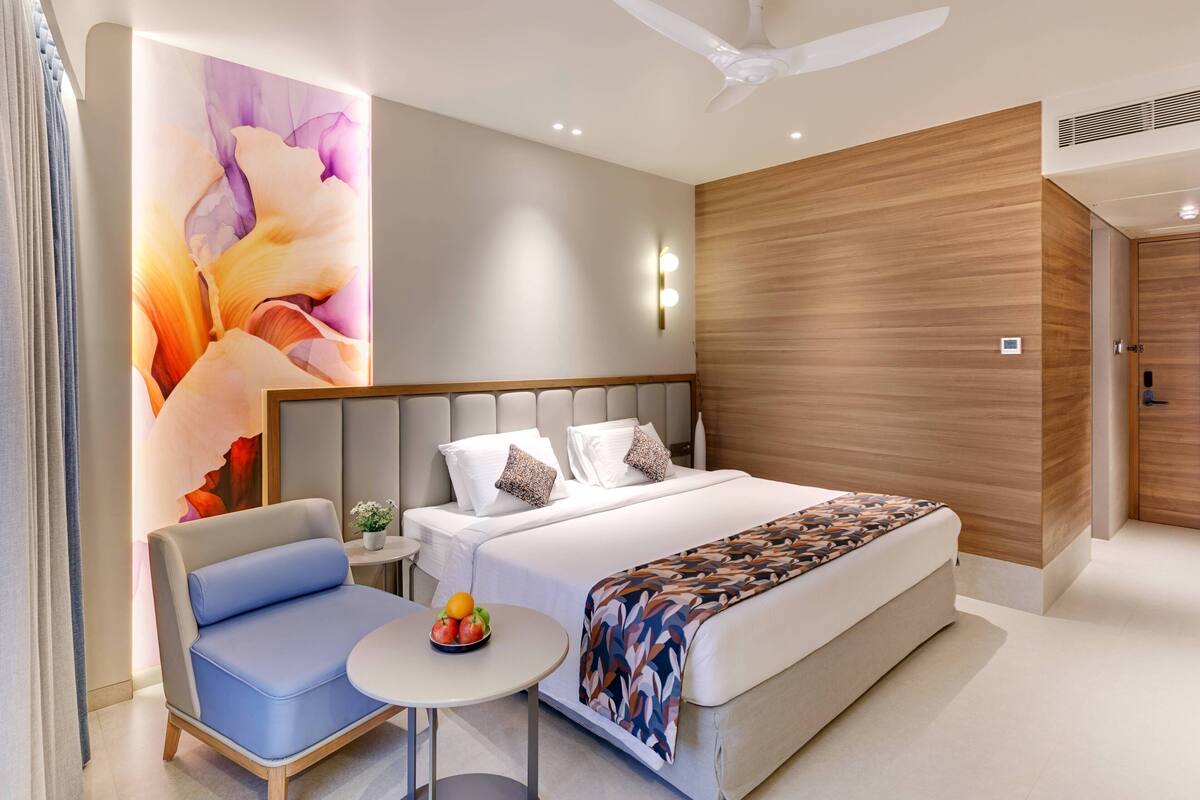 A modern hotel room with a large bed, a small round table with fruit, a blue armchair, and a decorative wall mural.