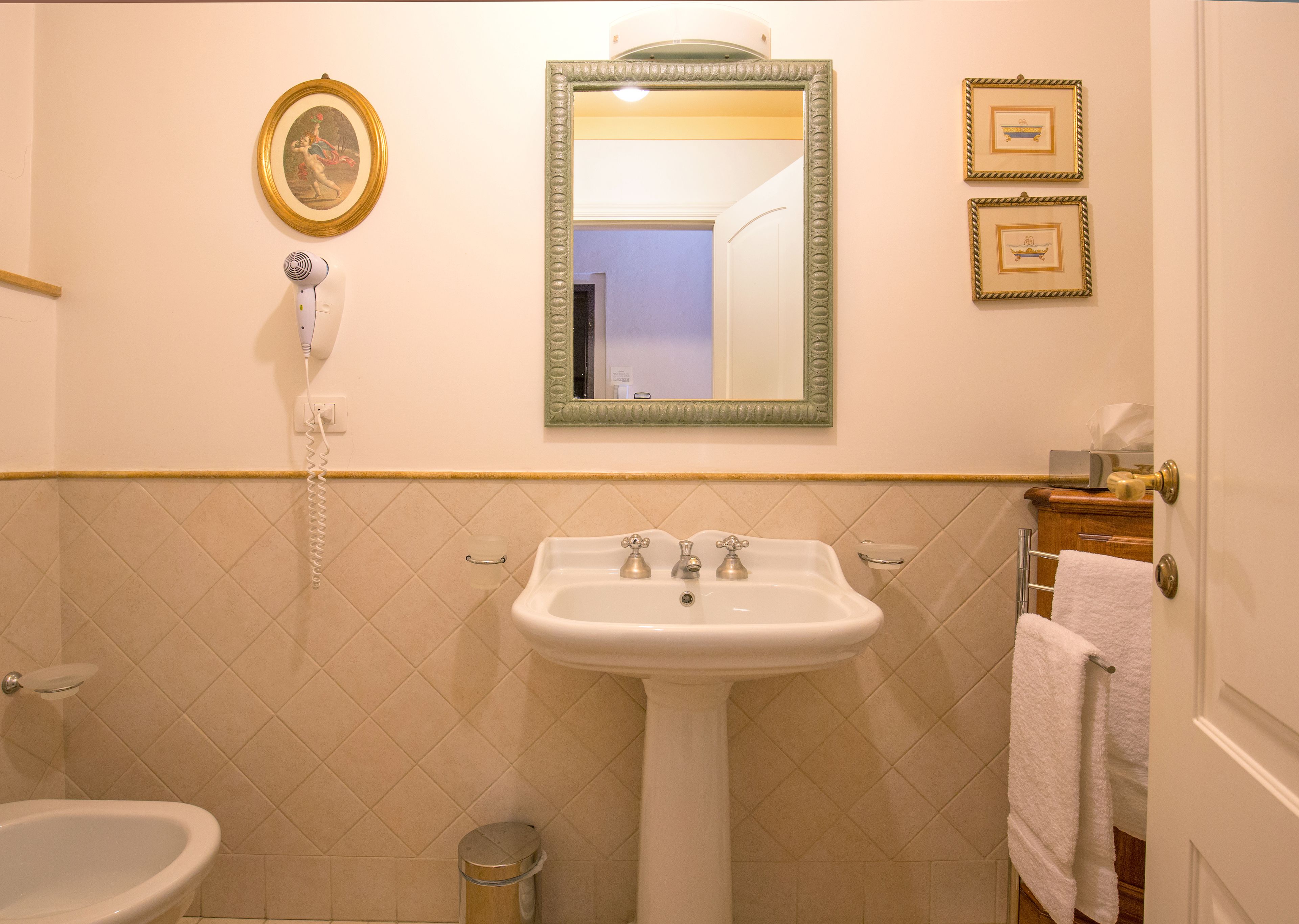 Een badkamer met een wastafel op voet, een toilet en een bidet. Boven de wastafel hangt een lijst met spiegel en aan de muur een fotolijst.