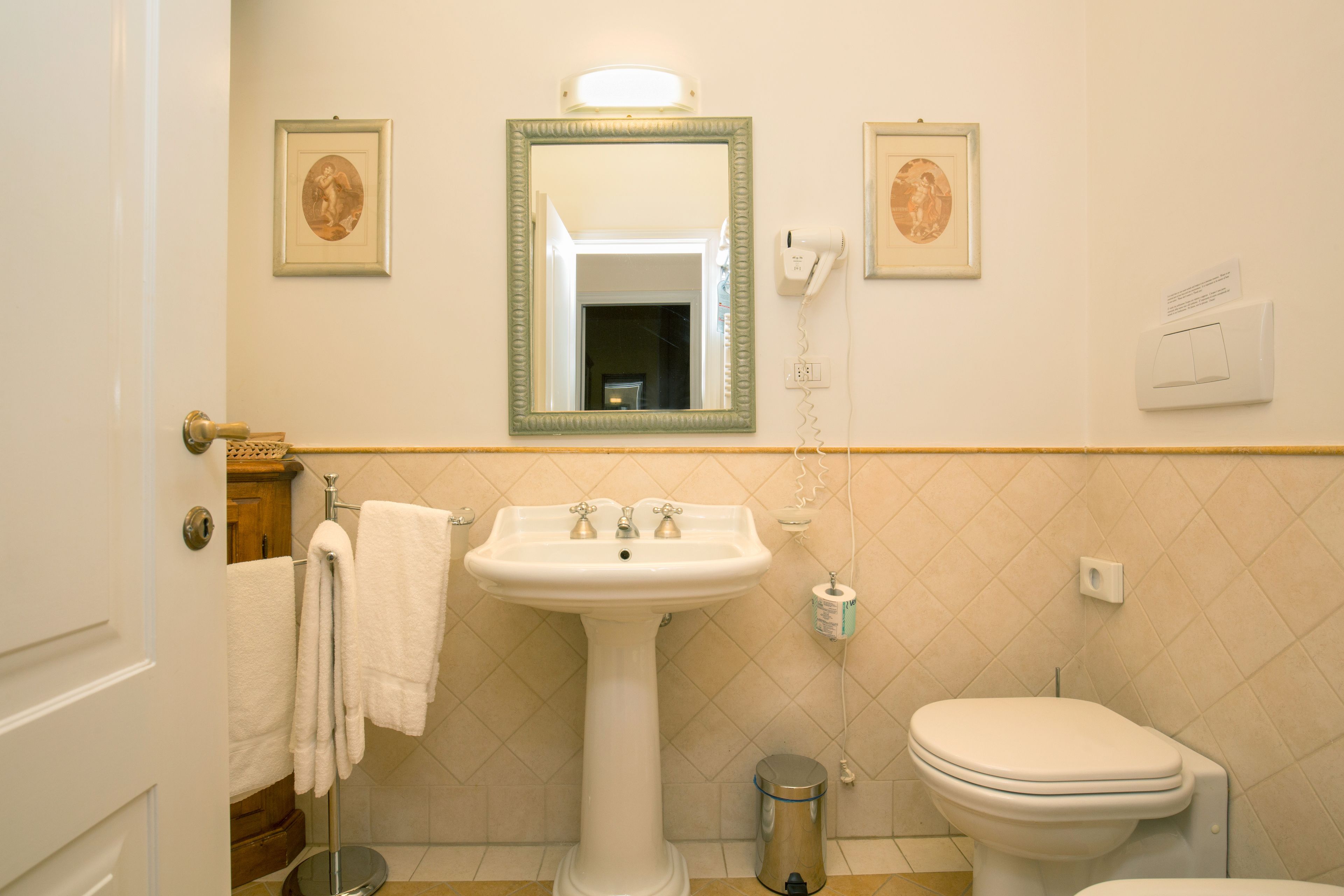 A bathroom with a pedestal sink, a toilet, a trash can, and framed pictures on the wall.