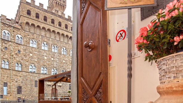 Eine Holztür mit Schild, ein großer Blumentopf und Blick auf ein historisches Gebäude.