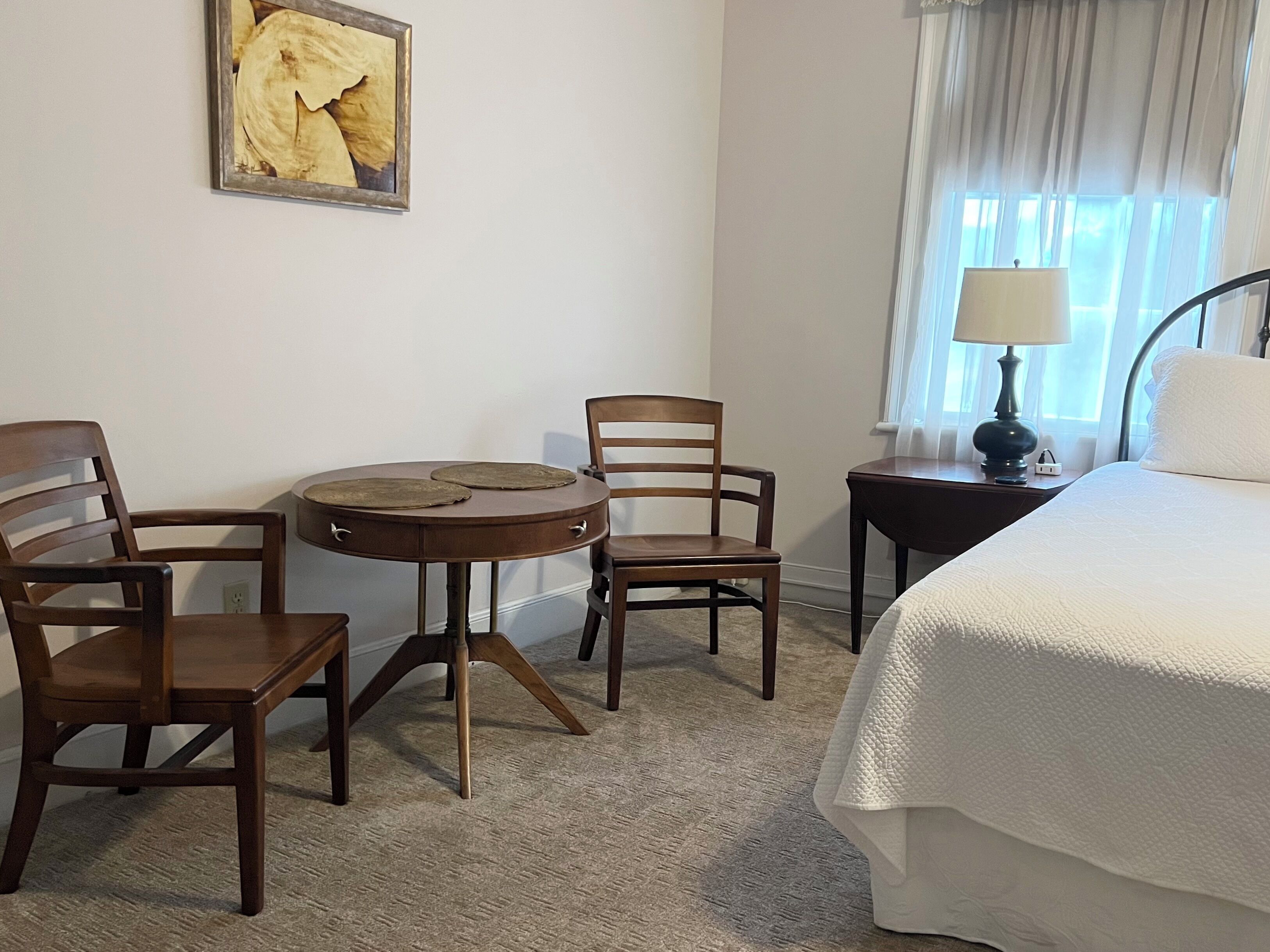 A hotel room with a bed, two wooden chairs, a small round table, a lamp, and a painting on the wall.