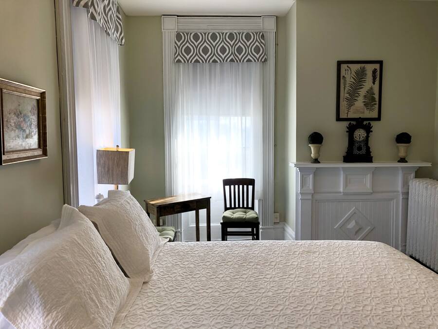 Un dormitorio con una cama grande, una silla, una mesita, una lámpara, una chimenea e impresión enmarcada de motivos botánicos en la pared.