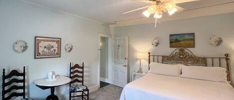 A bedroom with a bed, two chairs, a small table, a ceiling fan, and wall decorations.