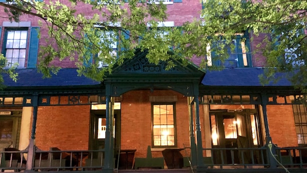 A building with a brick facade, a porch with a green railing, and a flower garden in front. - Rogues' Harbor Inn (Lansing)