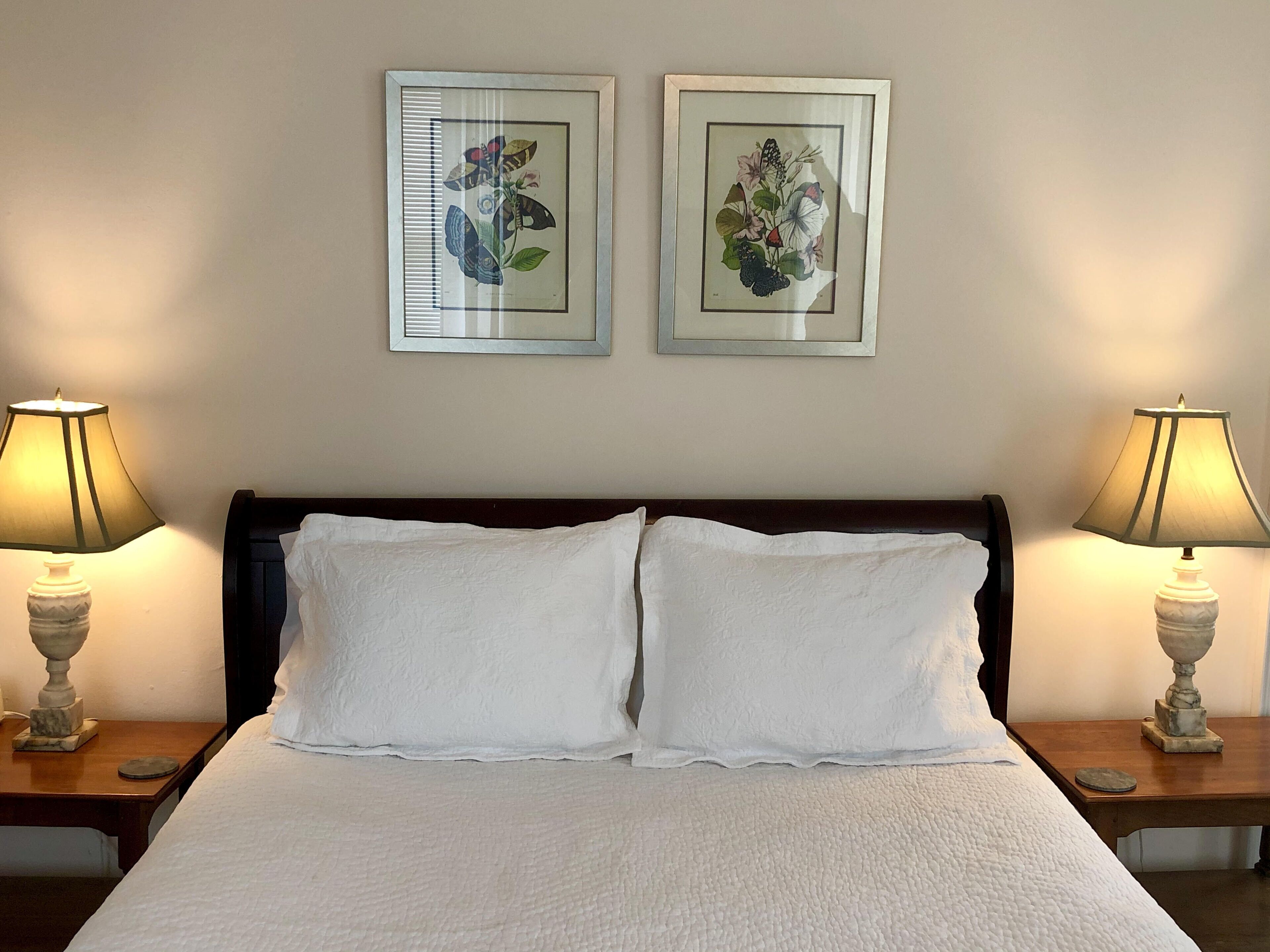 A neatly made bed with white linens, flanked by two bedside tables with lamps, and two framed botanical prints on the wall.