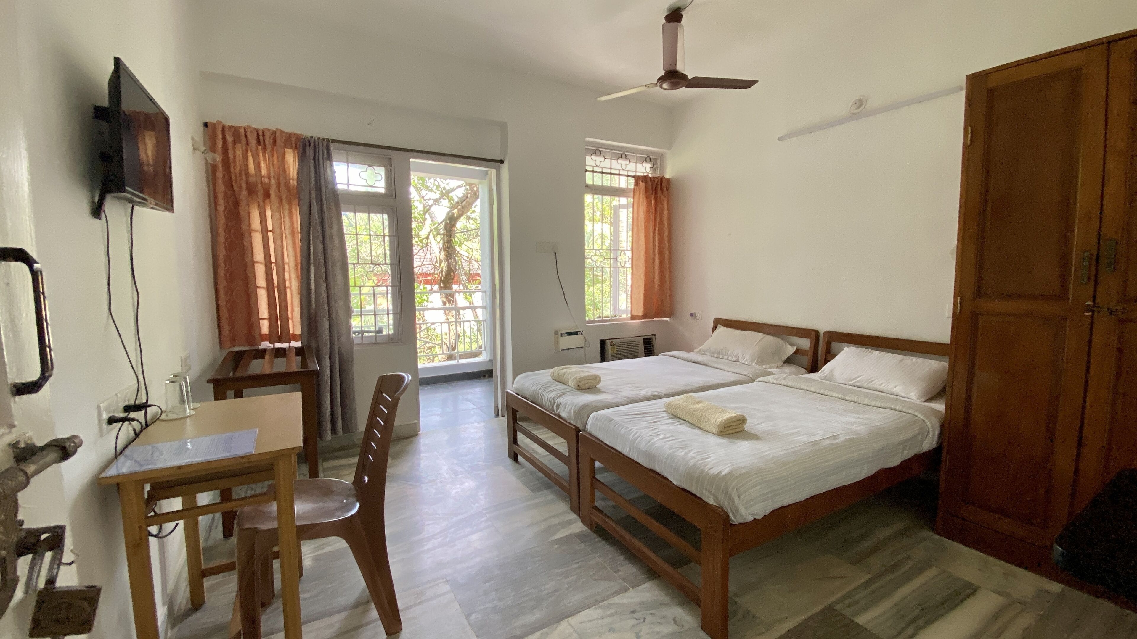 A room with two beds, a desk, a chair, a television, and a ceiling fan.