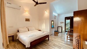 Un dormitorio con cama, ventilador de techo, equipo de aire acondicionado, una mesita y una puerta que da a otra habitación.