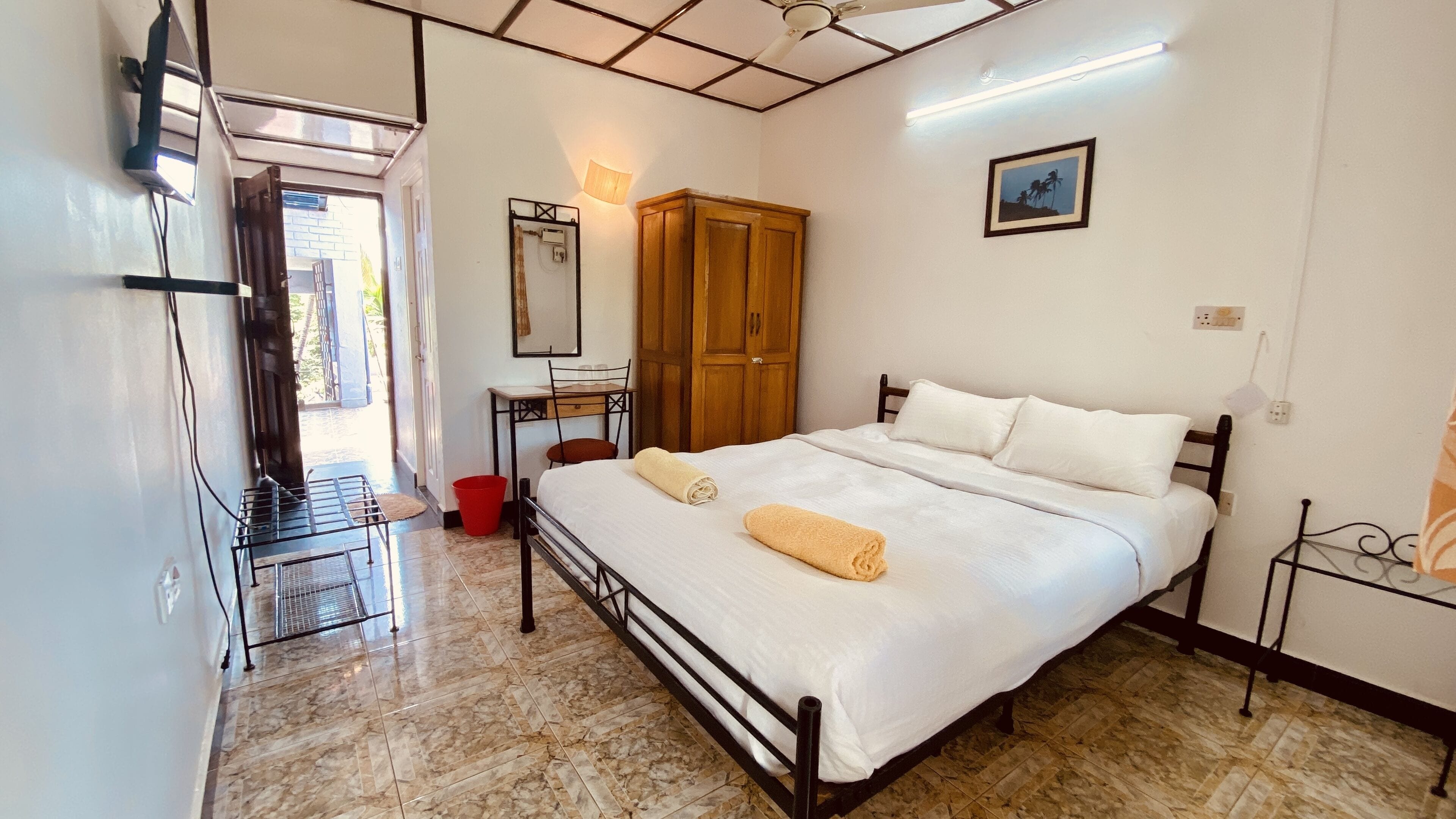 A hotel room with a bed, a wooden wardrobe, a desk with a chair, a red bucket, and a ceiling fan.