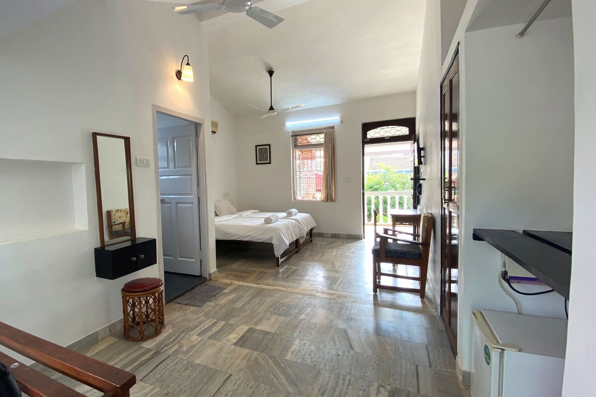 A hotel room with a bed, a desk, a chair, a small stool, a mirror, a ceiling fan, and a window with a view.