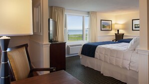 Een hotelkamer met een bed, bureau, stoel, televisie, raam met uitzicht op zee en een lamp.