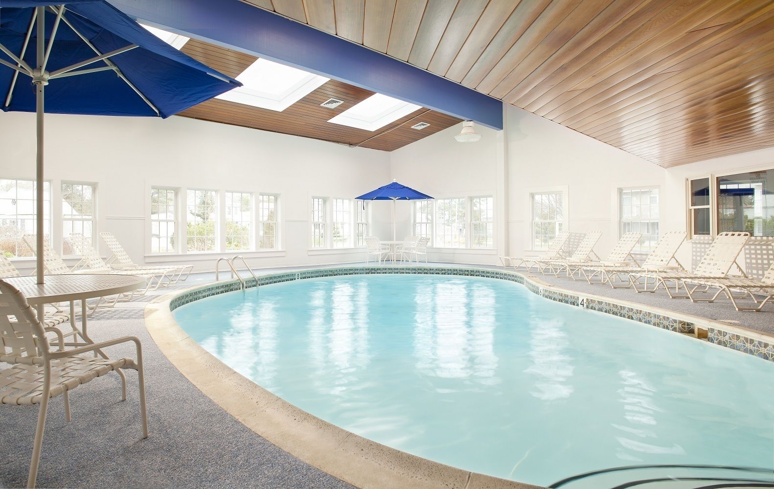 Una piscina interior con agua cristalina y azul, rodeada de sillones blancos y una sombrilla azul.