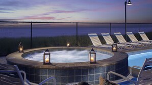 Een hottub met lounge fauteuils en kaarsen, omringd door een hek en met uitzicht op een wateroppervlak.