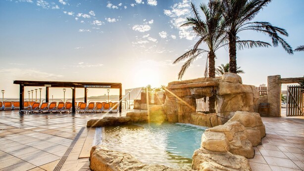 A pool area with a rock formation, palm trees, and orange chairs. - AX Sunny Coast Resort & Spa (St. Paul's Bay)