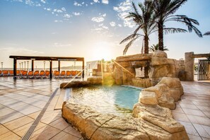 A pool area with a rock formation, palm trees, and orange chairs. - AX Sunny Coast Resort & Spa (St. Paul's Bay)