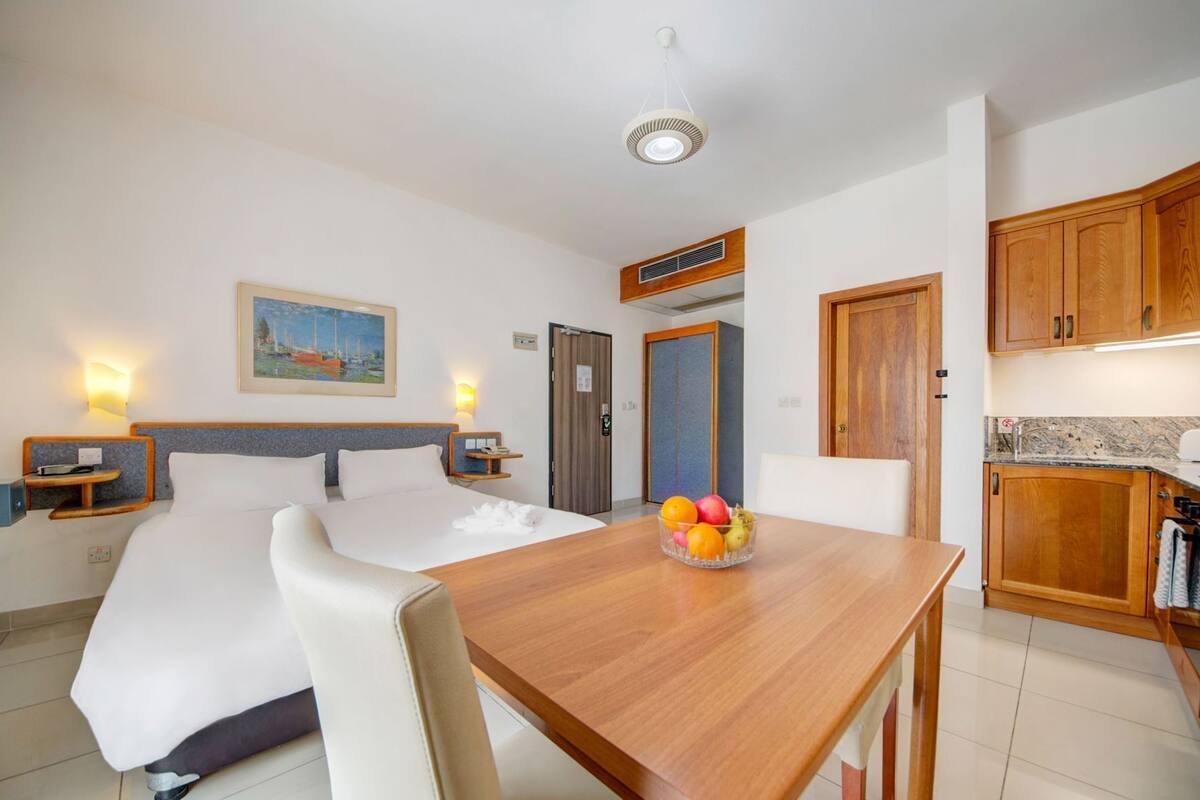 A hotel room with a bed, bedside tables, a desk with a fruit bowl, a painting, and a kitchenette.
