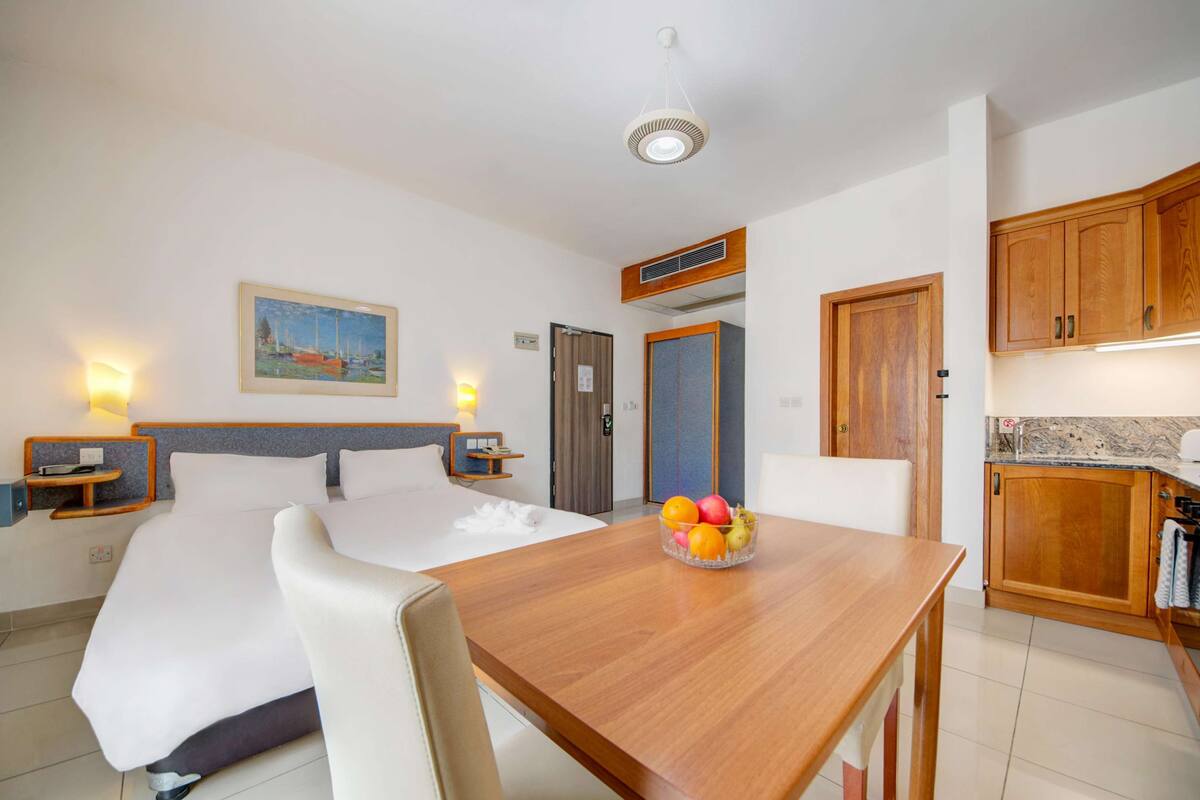 A hotel room with a bed, bedside tables, a wooden dining table with fruit, a painting on the wall, and wooden cabinets.