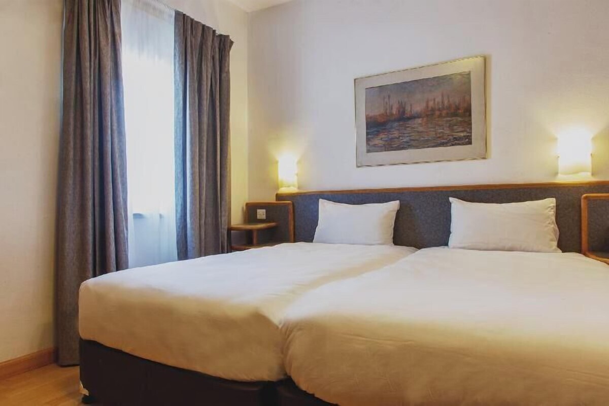 A double bed with a headboard, two bedside tables with lamps, a painting on the wall, and a window with curtains.