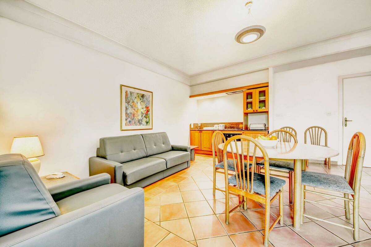 A living room with a grey sofa, a dining table with chairs, a kitchen area, and a painting on the wall.