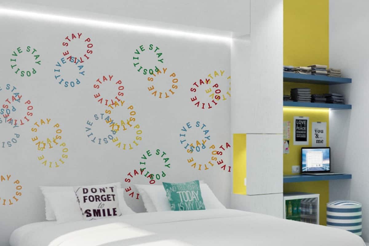 Ein modernes Schlafzimmer mit einem weißen Bett, einer Wand mit motivierenden Sprüchen, einem Schreibtisch mit einem Laptop und einem Bücherregal.