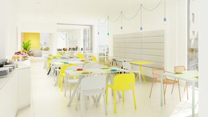 Ein heller, moderner Essbereich mit weißen und gelben Tischen und Stühlen, im Hintergrund eine Küche und Pendelleuchten.