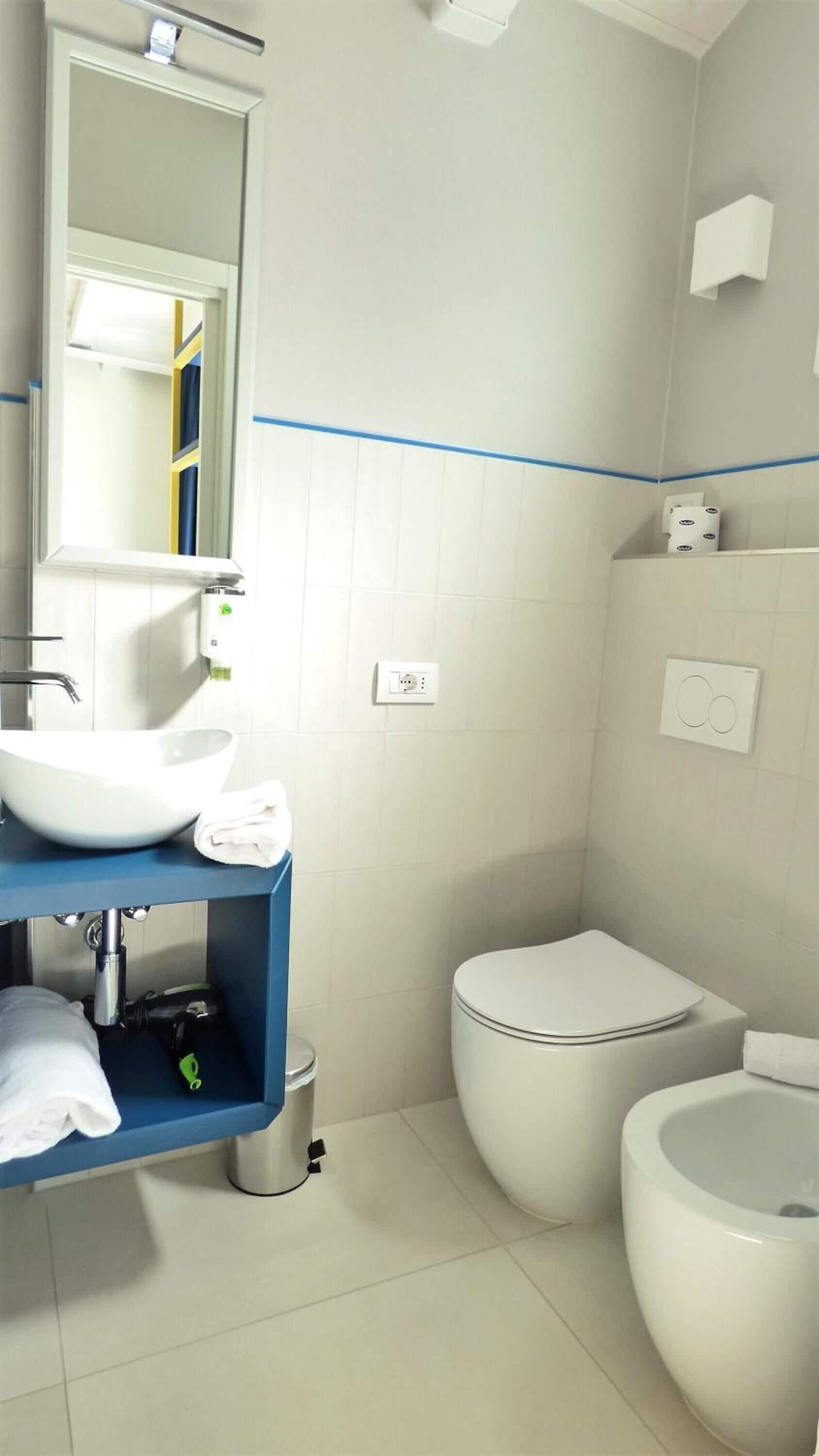 Ein modernes Badezimmer mit einem weißen Waschbecken, Spiegel und Toilette. Darunter befindet sich ein blaues Regal und eine kleine Mülleimer.