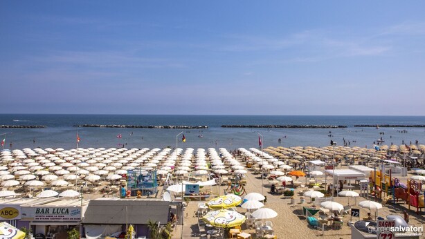 Ein Strand mit vielen Sonnenschirmen und Liegestühlen, eine Bar namens 'BAR LUCA' und Blick aufs Meer.
