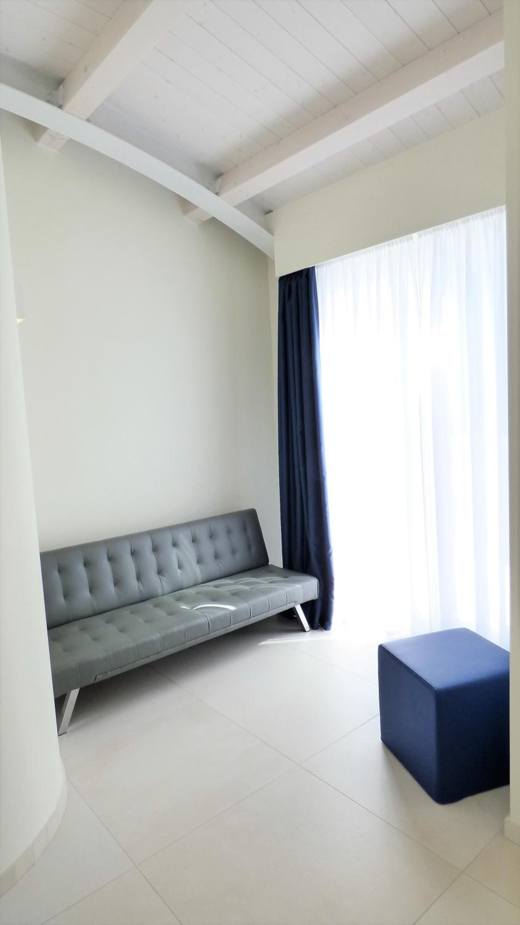 Una stanza moderna con un divano grigio, un poggiapiedi blu scuro e un'ampia finestra con tende leggere.