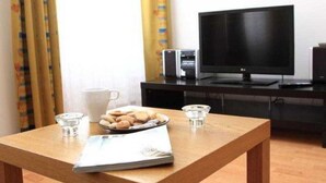 A room with a wooden table, a plate of cookies, two glasses, a white mug, and a black TV on a black stand. - Theatre Residence Apartments (Prague)
