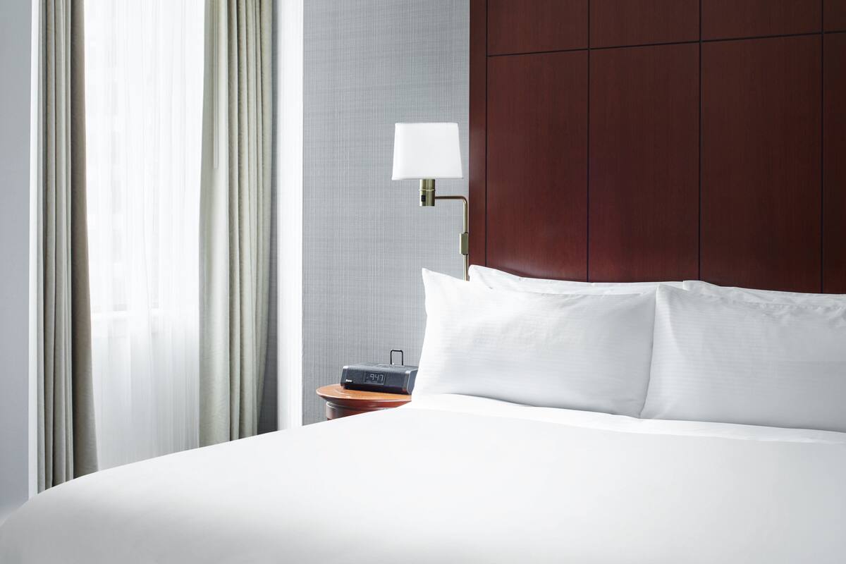 Ein Hotelzimmer mit einem ordentlich bezogenen Bett, einem Nachttisch mit Lampe und einer digitalen Uhr.