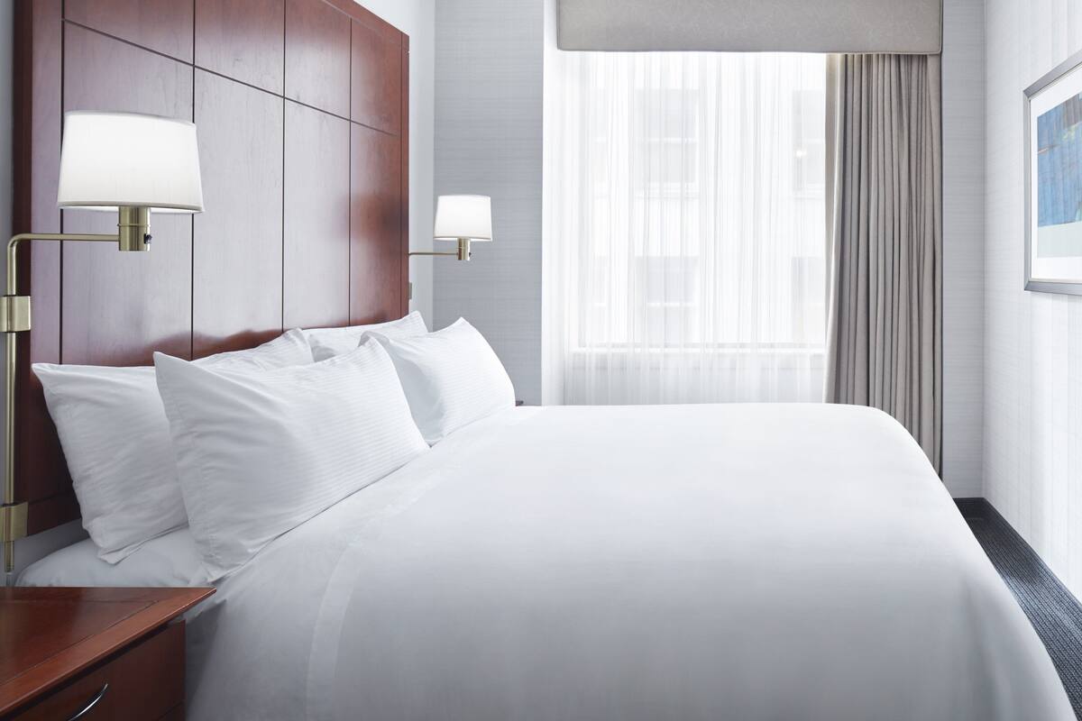 Ein Hotelzimmer mit einem großen Bett, zwei Nachttischlampen, einem hölzernen Kopfteil, einem Fenster mit Vorhängen und einem gerahmten Bild an der Wand.